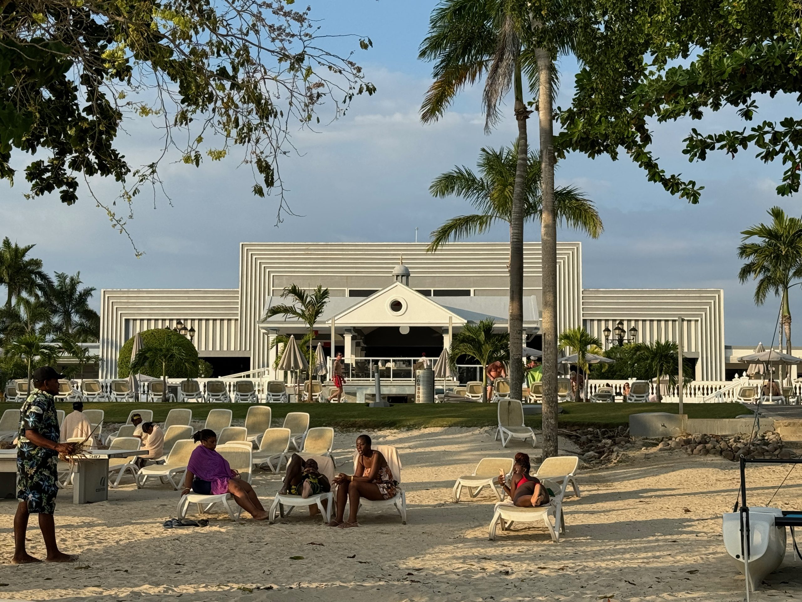 People relaxing on lounge chairs on a beach in Negril, Jamaica, with palm trees and a resort building behind them.